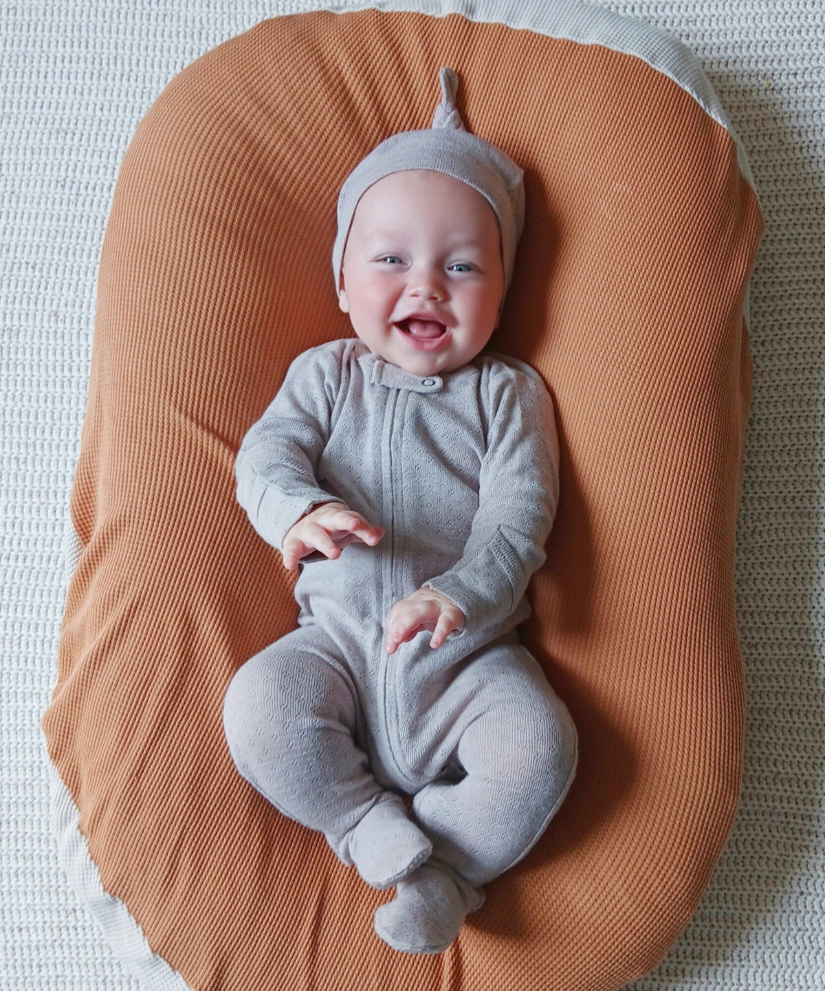 Baby in a gray outfit sitting on an orange cushion with a white background