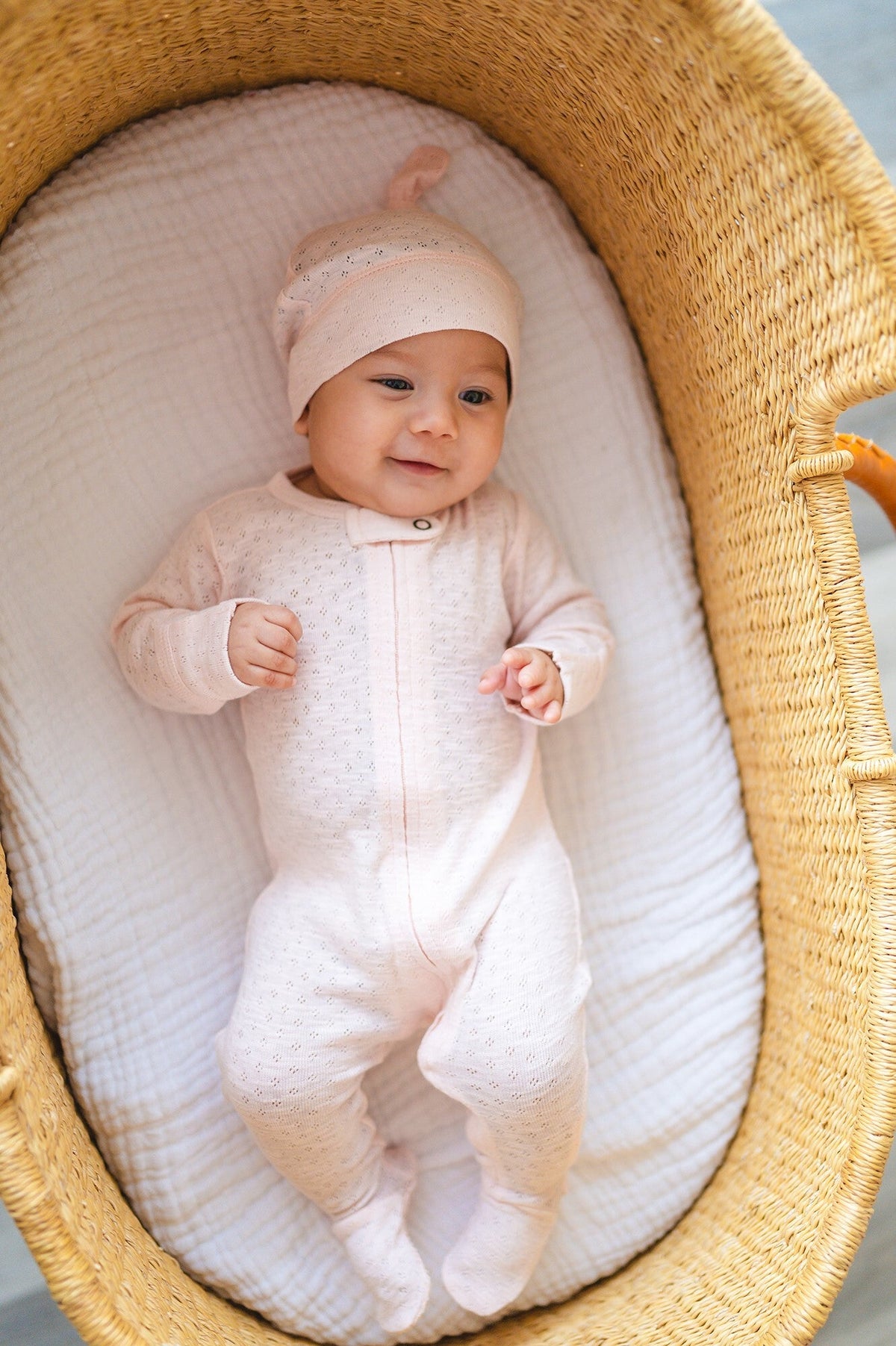 Baby in a white outfit and pink hat lying in a wicker basket with white bedding.