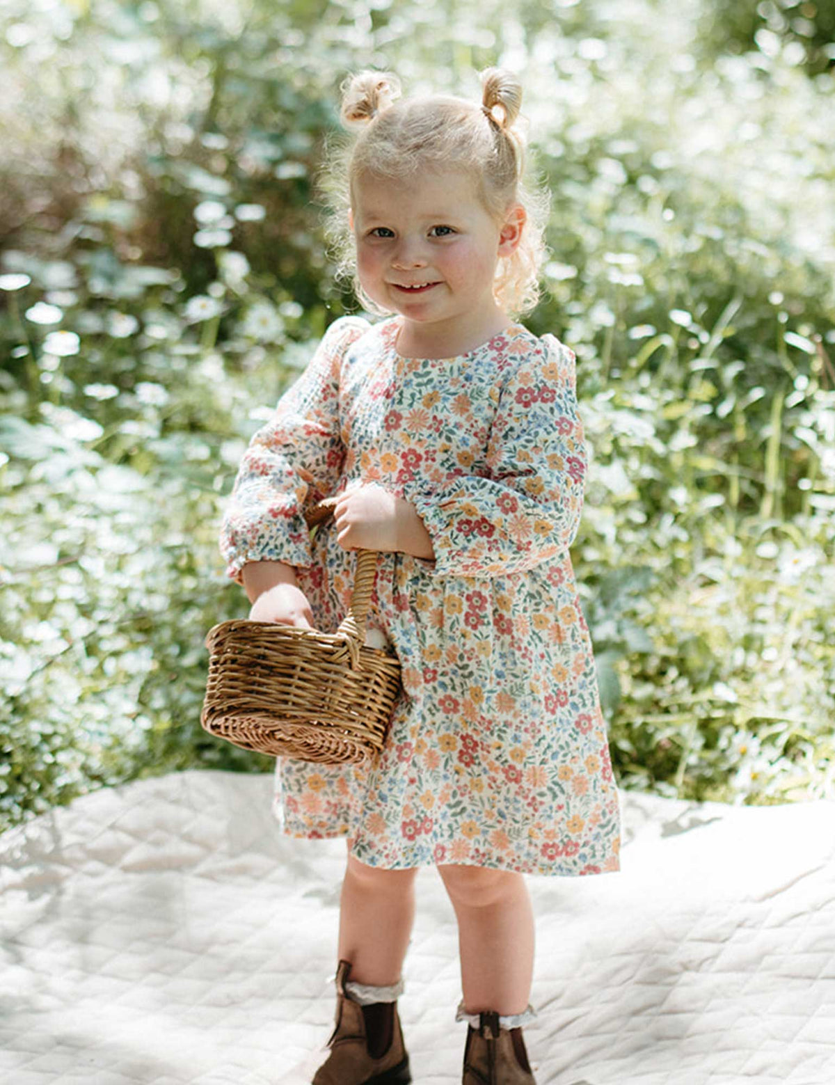 Young girl in a floral dress holding a basket outdoors with greenery in the background