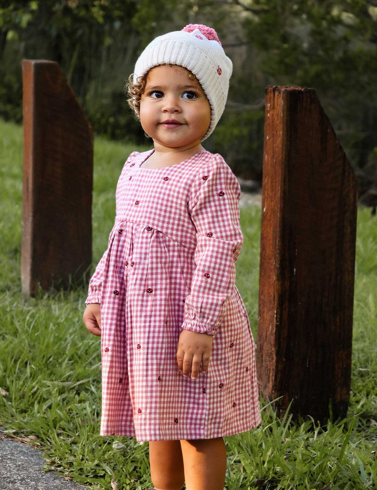 Child wearing a pink checkered dress and white hat with a pink bow outdoors.
