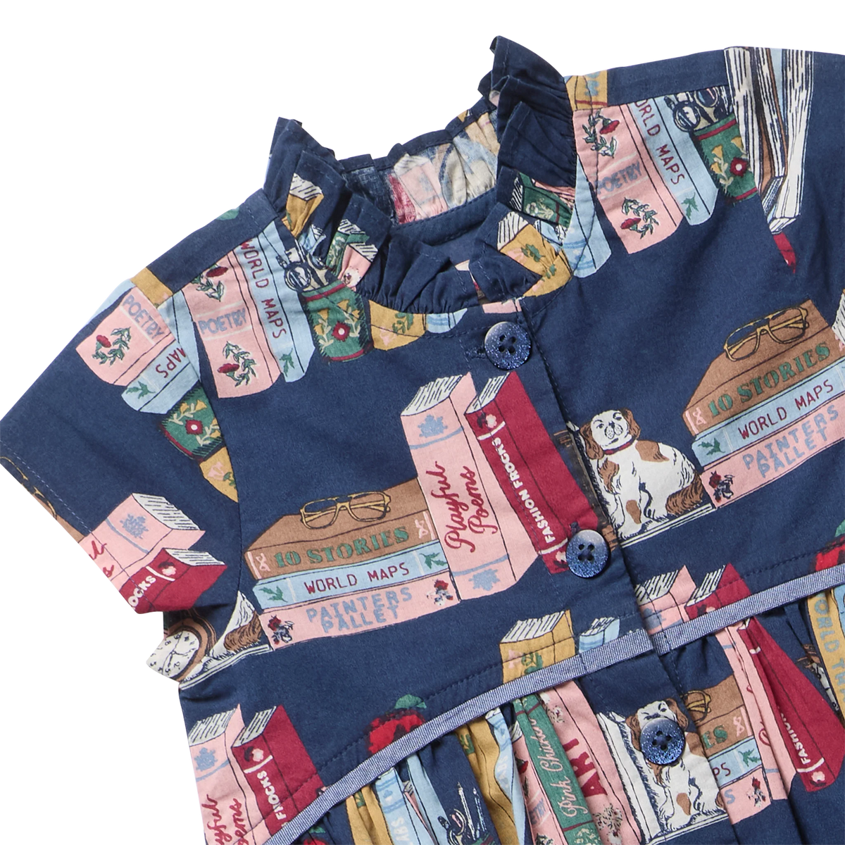 A close-up of a navy blue short-sleeve dress featuring a whimsical book-print pattern with illustrations of books, a cat, and text like 'Fairy Tales' and 'World Maps'. The dress has a ruffled mock neck, blue buttons, and a tiered skirt.