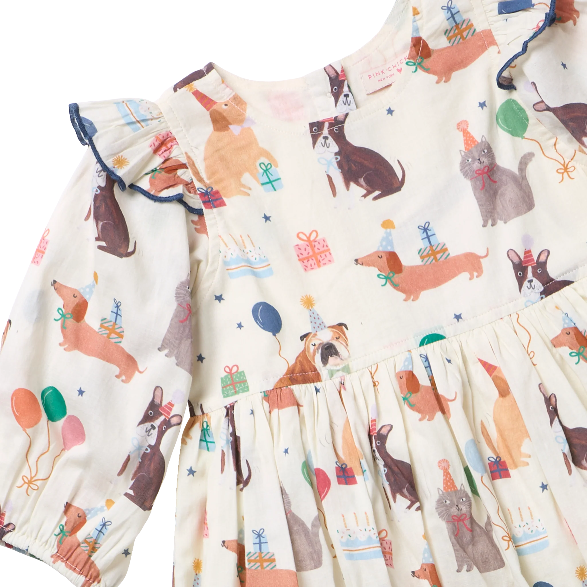 A close-up of a baby dress with a cream-colored base and an all-over print of dachshunds, birthday candles, party hats, balloons, and gift boxes. The dress features ruffled long sleeves with dark blue trim and a ruffled bodice and skirt.