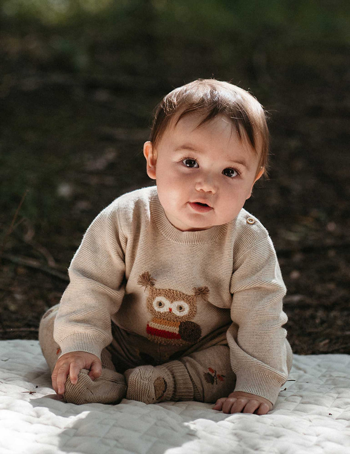 Baby wearing a beige sweater with an owl design, sitting on a blanket outdoors.