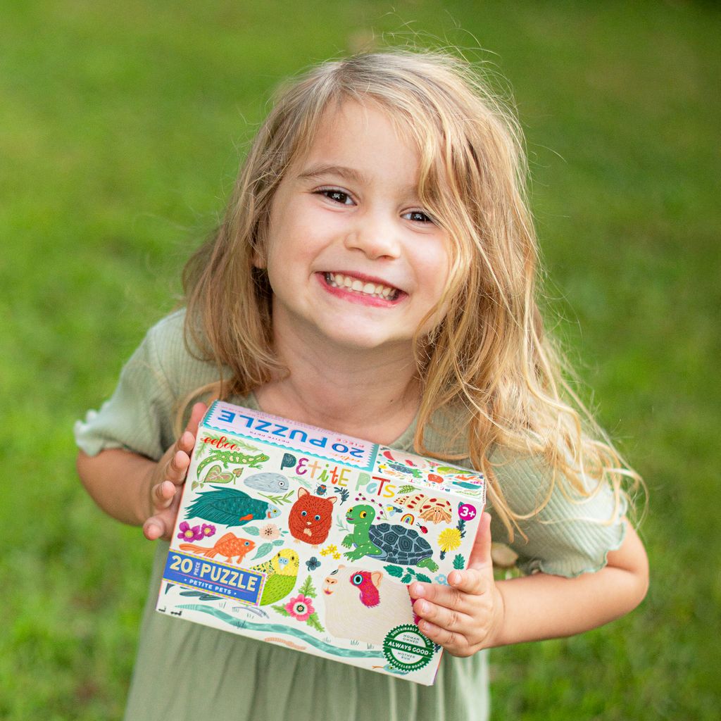 A young smiling girl outdoors in a green setting, happily holding up the Eeboo Petite Pets 20-Piece Jigsaw Puzzle box.