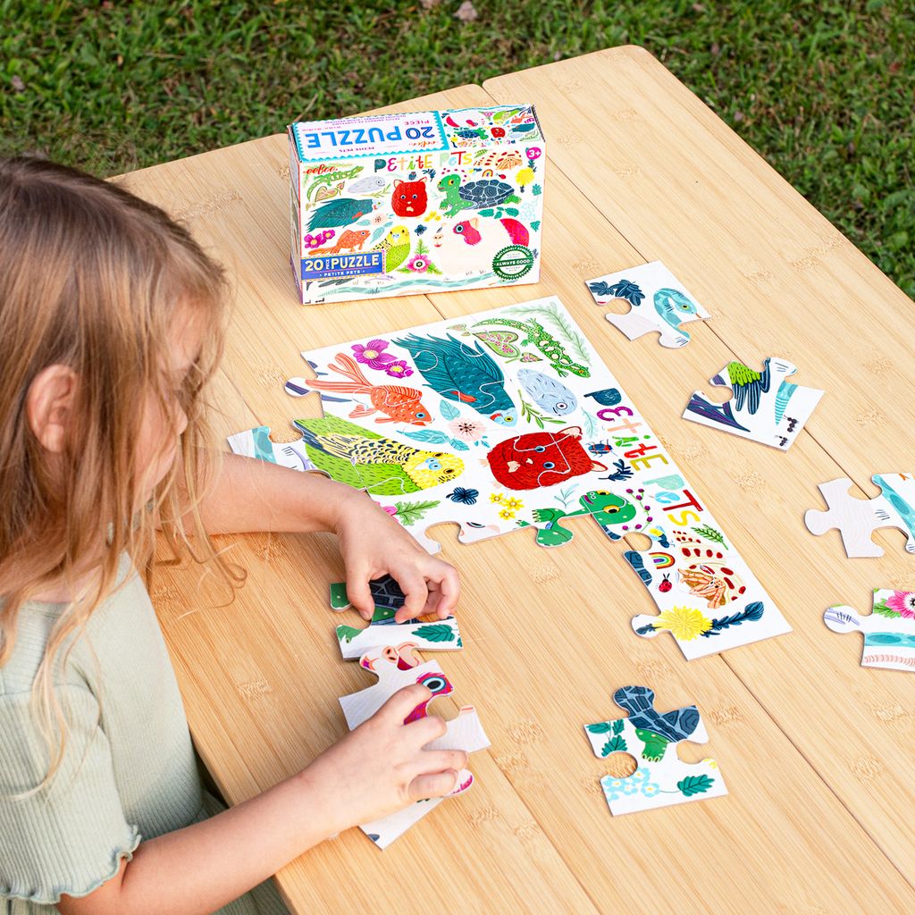 A young girl sitting outdoors at a wooden table, working on putting together the Eeboo Petite Pets 20-Piece Jigsaw Puzzle with the box resting nearby.