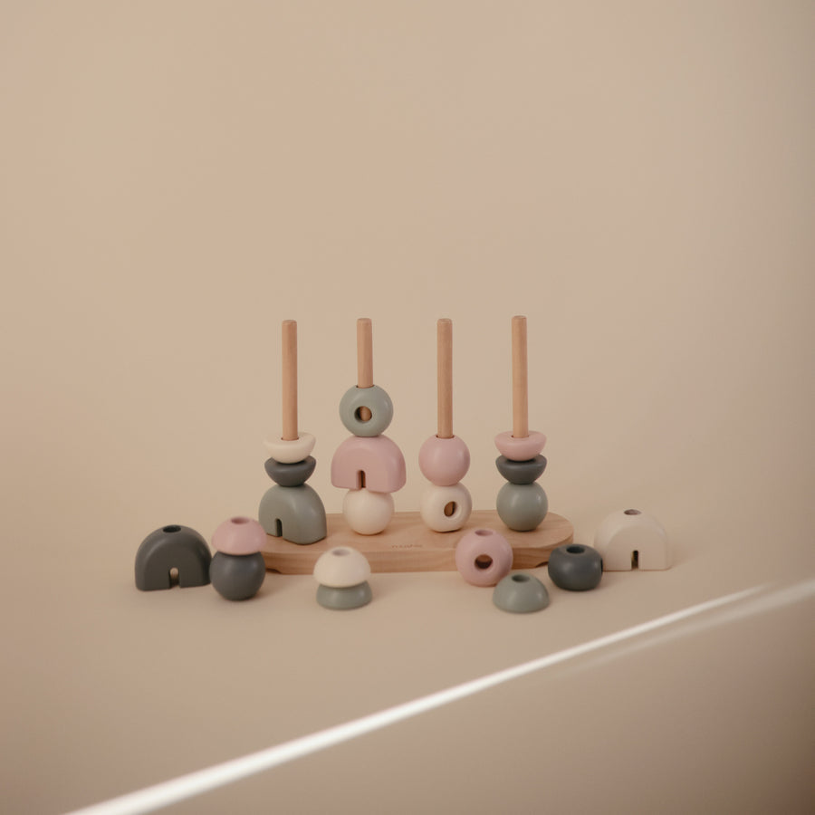A wooden multi-shape stacker toy by Mushie & Co featuring four vertical pegs on a wooden base, with additional geometric wooden pieces scattered around the base in soft tones of cream, pink, sage green, and charcoal gray.