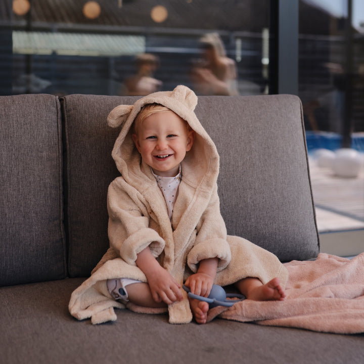Child in a beige robe with a hood sitting on a couch indoors.