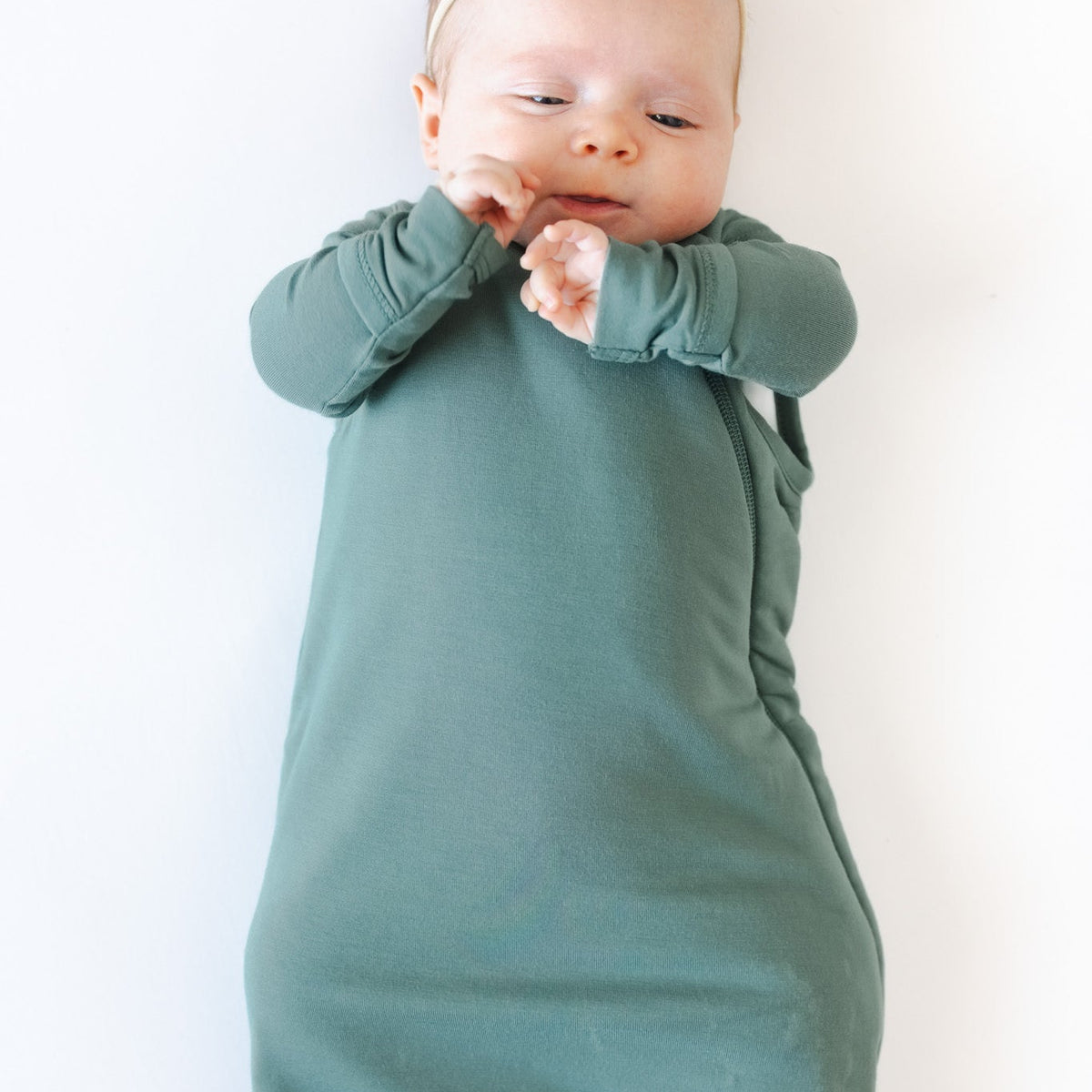 A baby wearing a muted sage green color long-sleeve wrap swaddle and a matching bow headband, lying on a plain white background.