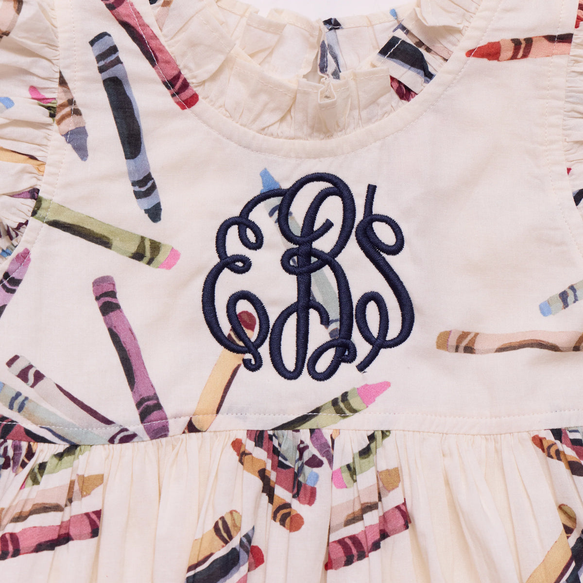 Close-up of a cream-colored children's dress with a colorful crayon print and a navy blue monogram embroidered on the chest.