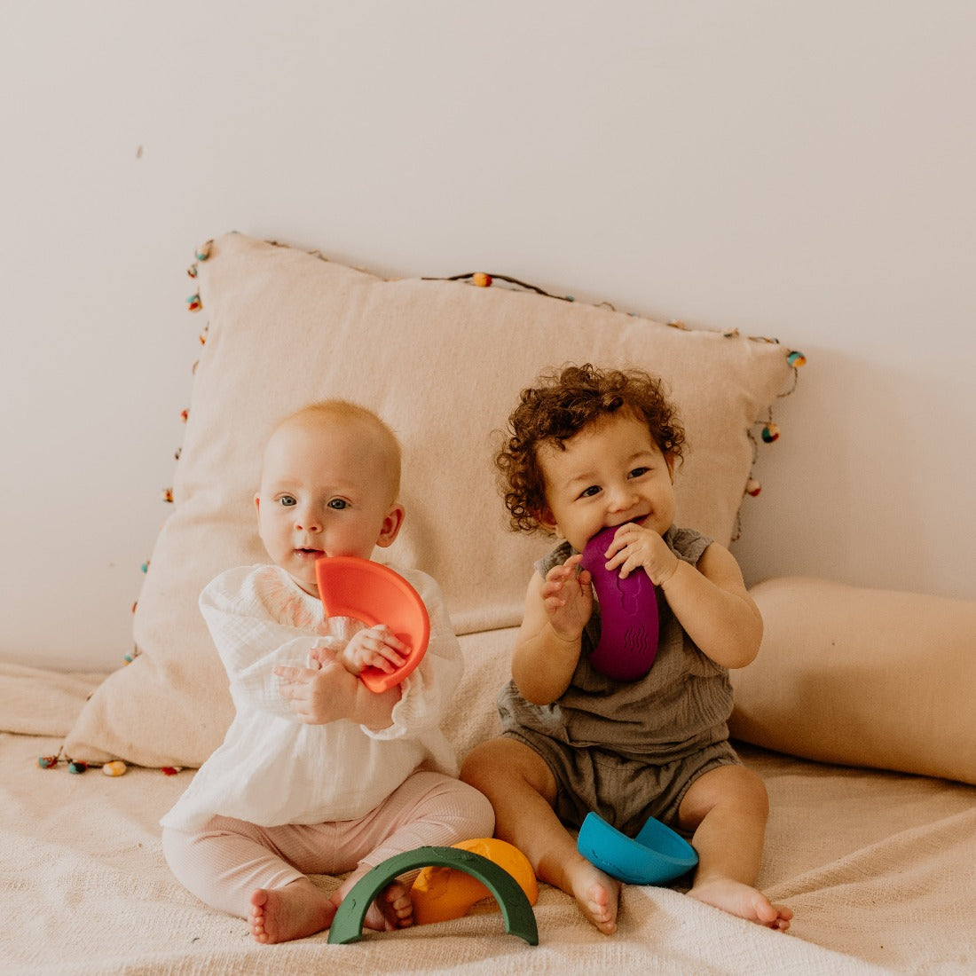 Two babies sitting on a couch with toys, one holding an orange toy and the other a purple toy.