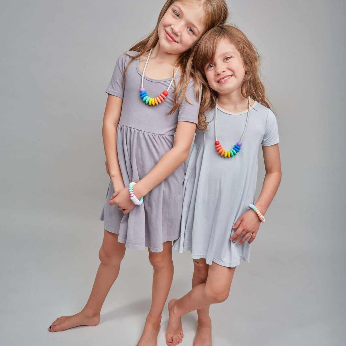 Two young girls wearing gray dresses with colorful necklaces on a plain background