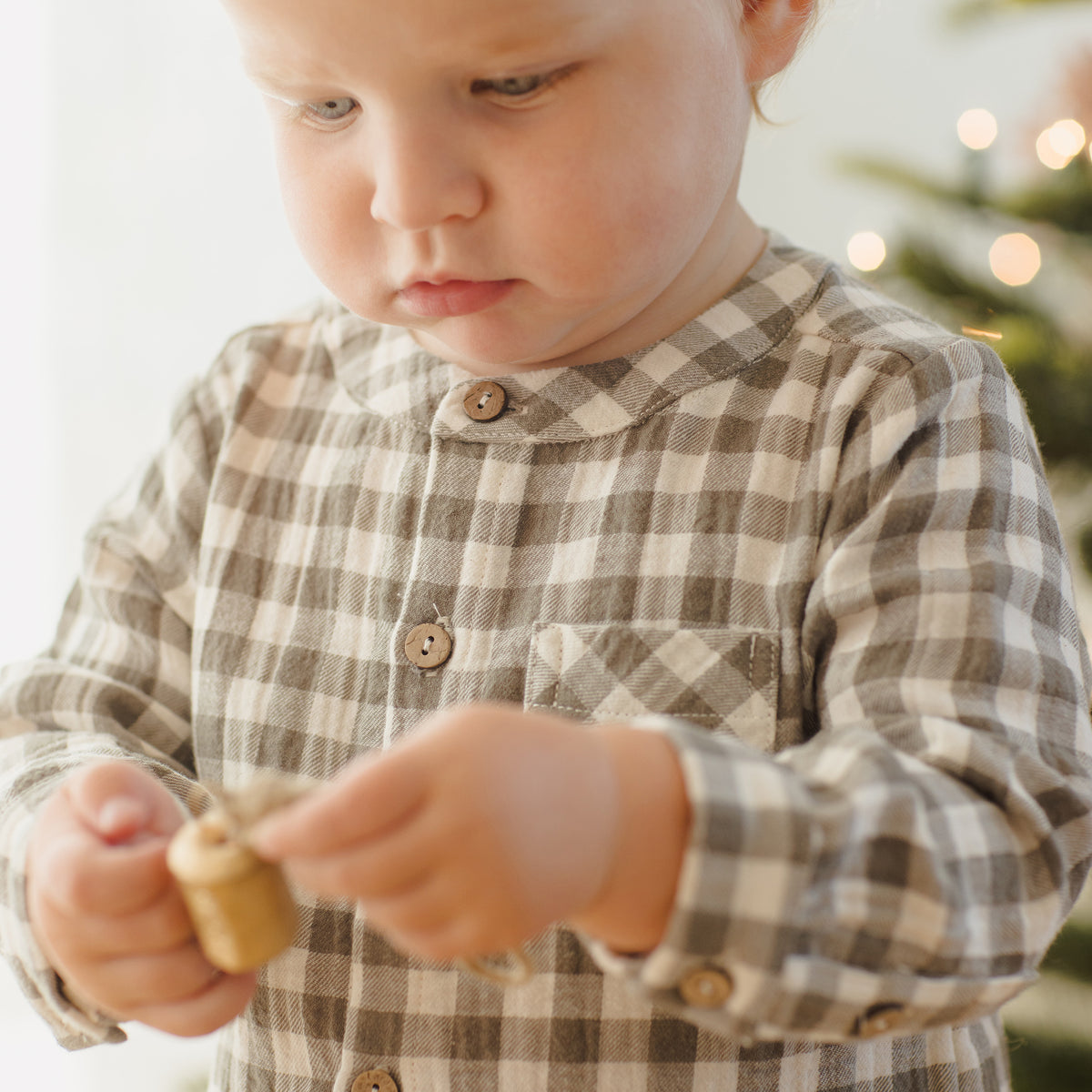 Child wearing a plaid shirt holding a small gold ornament with a blurred Christmas tree in the background.