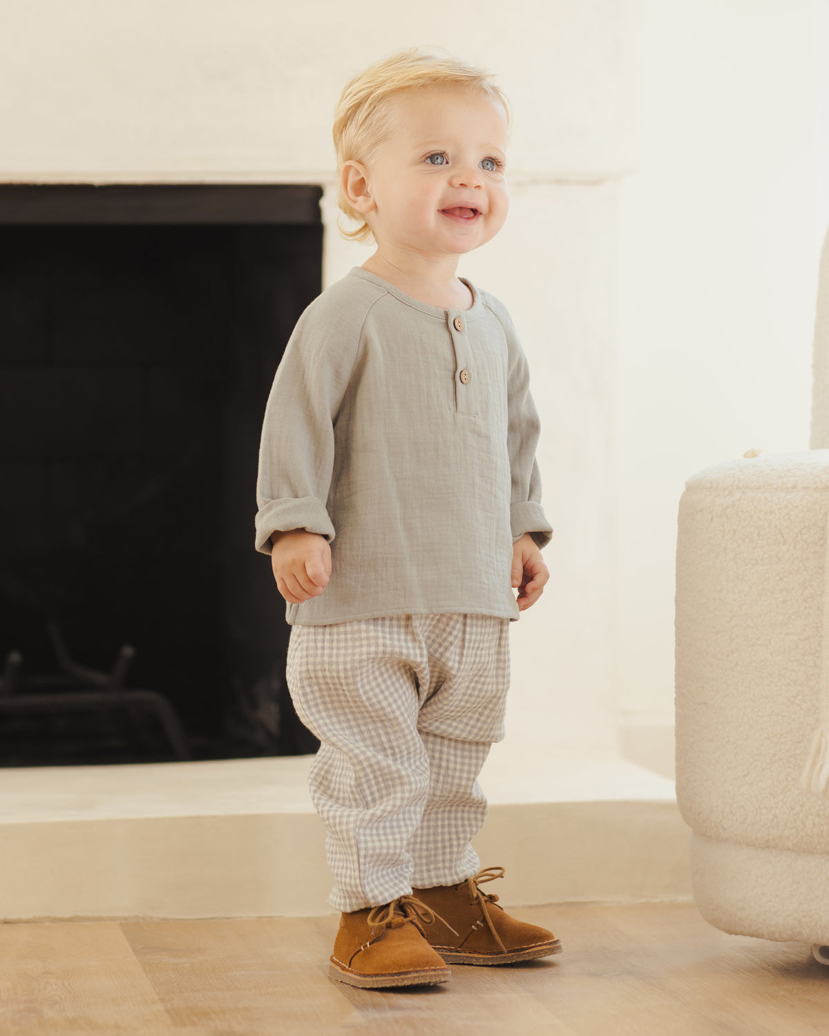 Child wearing a gray shirt and checkered pants standing in a room with a fireplace.