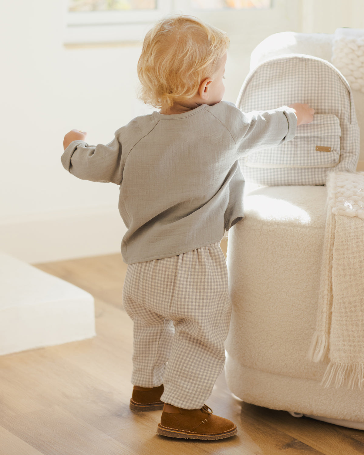 Child in a beige jacket and checkered pants standing near a white chair in a bright room.