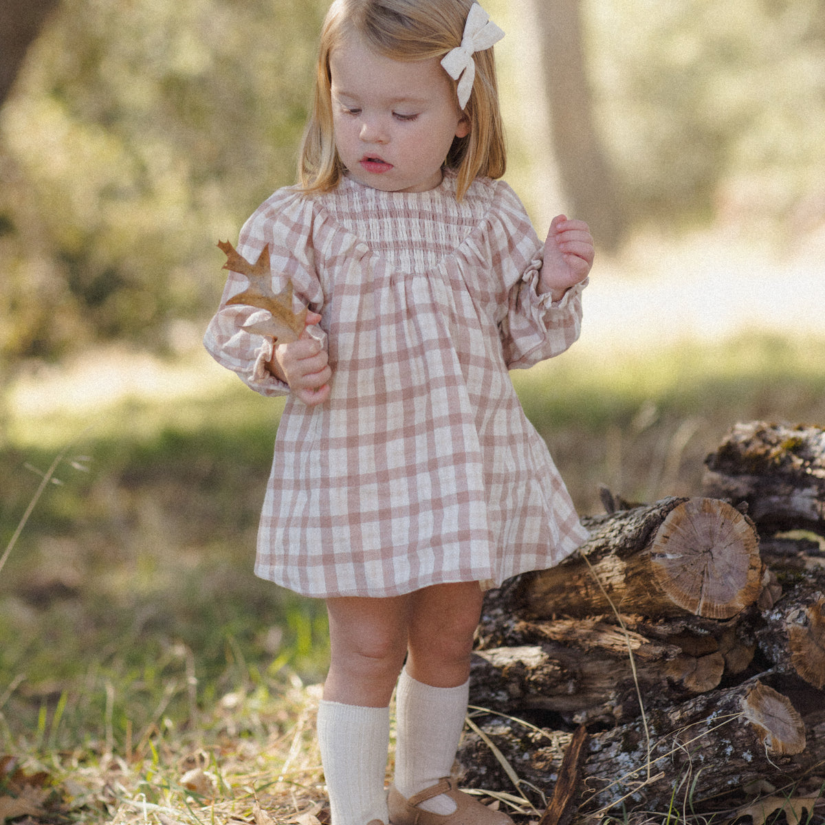 A young girl stands outdoors in a wooded setting wearing a pink and white gingham long-sleeve dress, cream knee-high socks, and tan shoes. She has a white bow in her hair and is standing near a log pile.