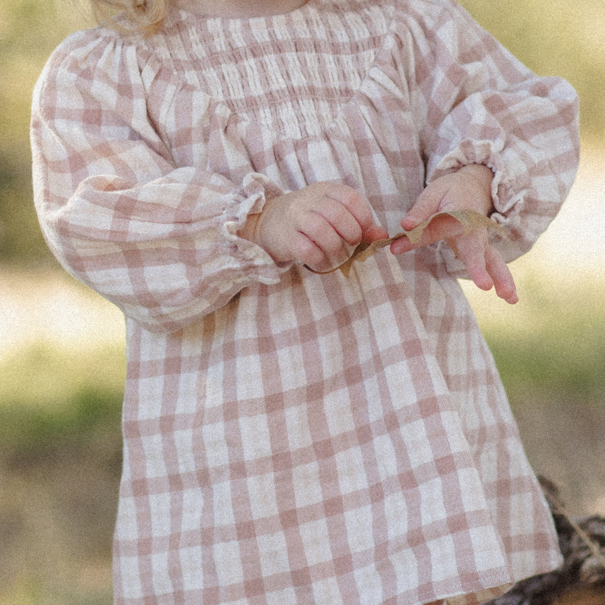 A toddler wearing a long-sleeve smocked blouse with a dusty pink and white gingham pattern, photographed outdoors with a blurred natural background.