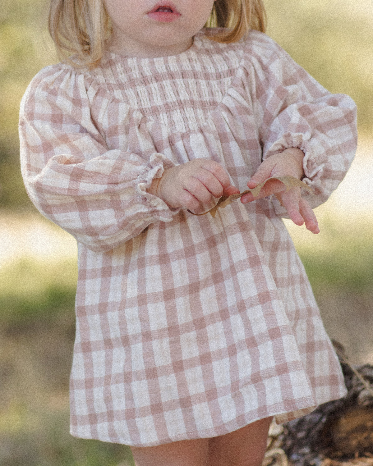 A toddler wearing a long-sleeve smocked blouse with a dusty pink and white gingham pattern, photographed outdoors with a blurred natural background.