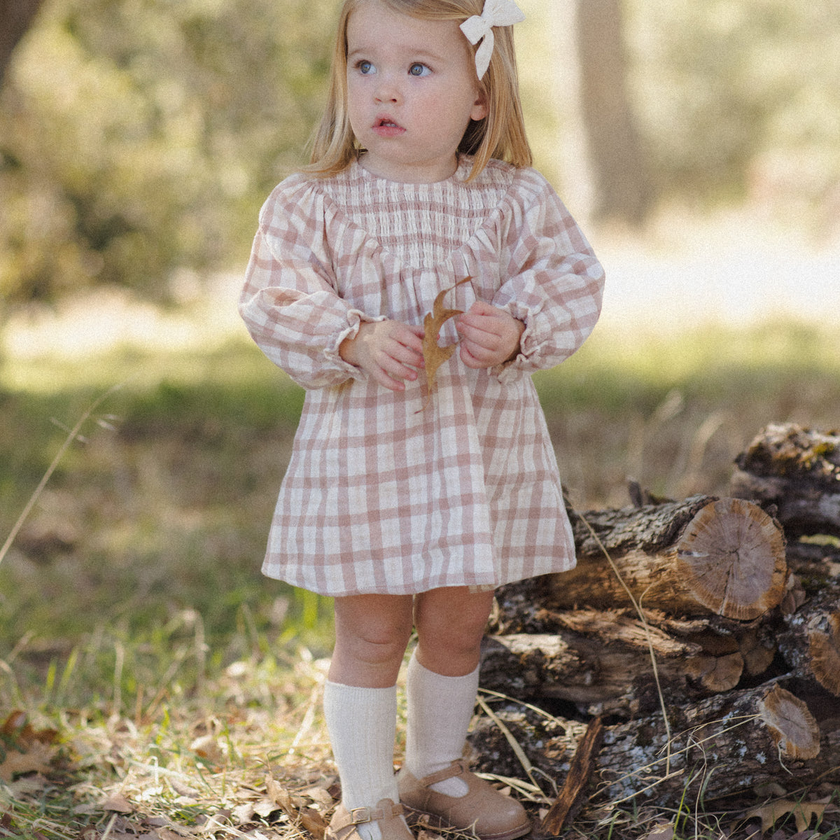 A young girl stands outdoors in a wooded setting wearing a pink and white gingham long-sleeve dress, white socks, and tan shoes, with a white bow in her hair.