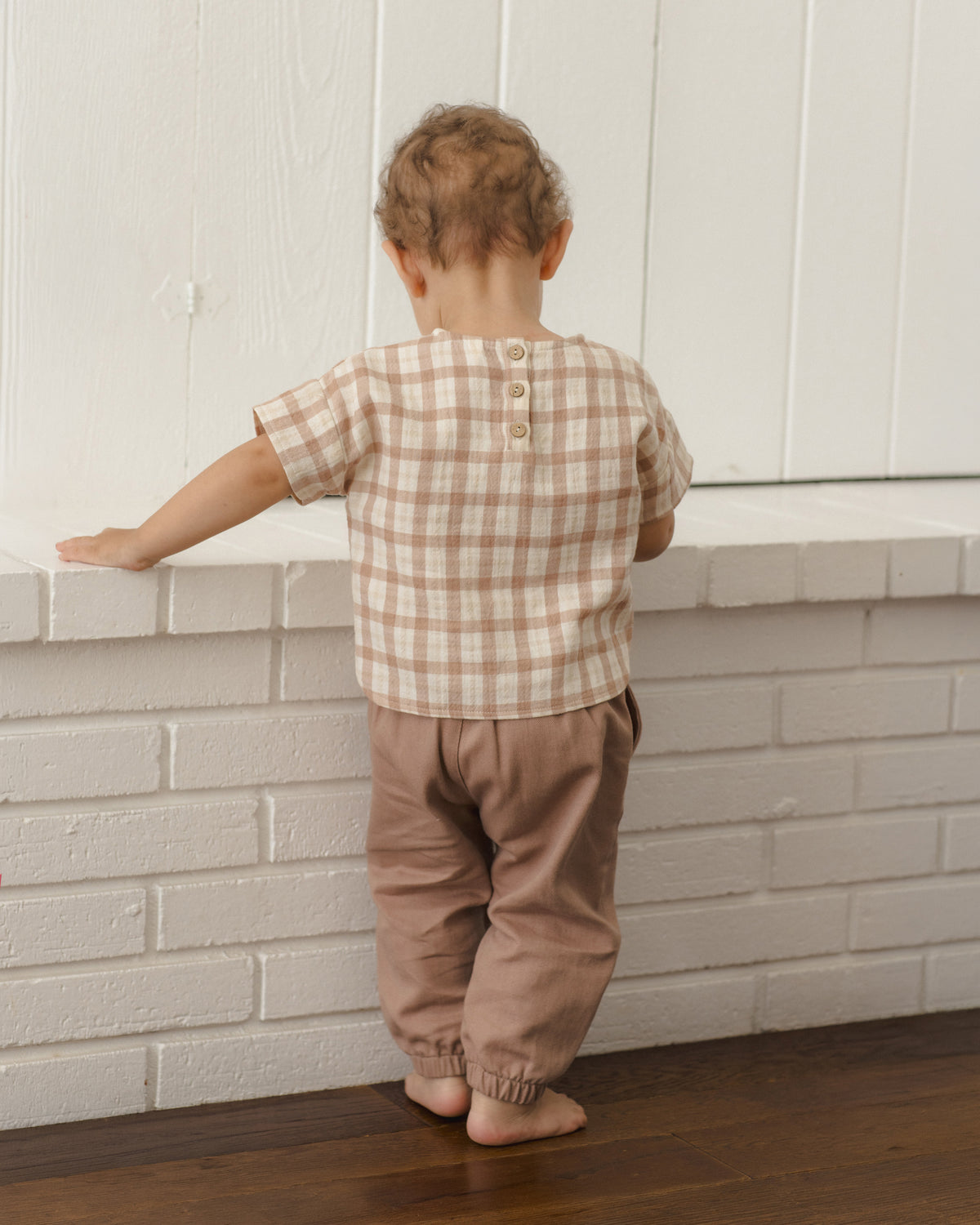 A toddler standing with their back to the camera, wearing a beige and brown gingham short-sleeve top with three buttons at the back and dusty brown pants, leaning on a white brick ledge.