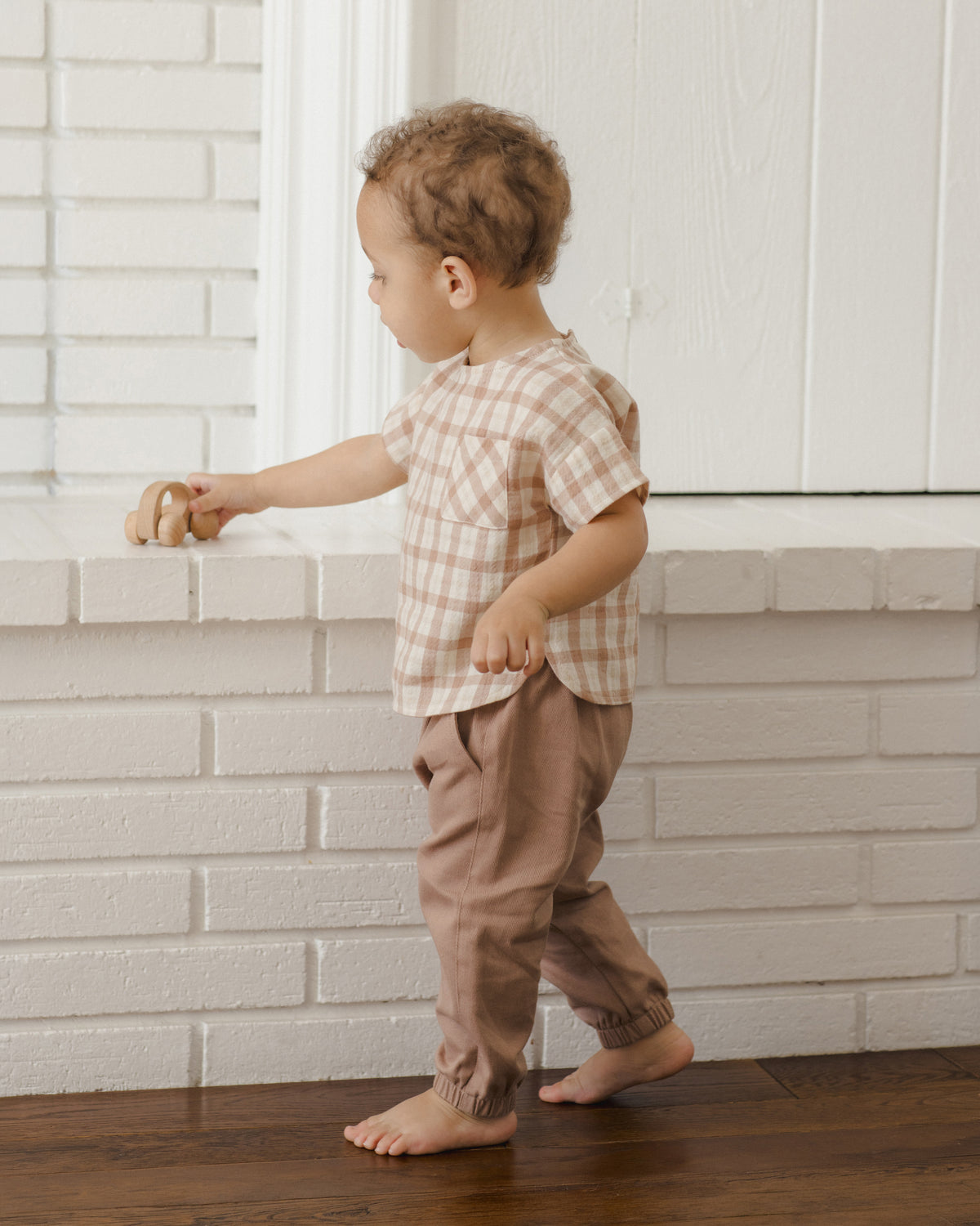 A toddler standing barefoot in front of a white brick wall, wearing a short-sleeve beige and white gingham shirt with brown pants.