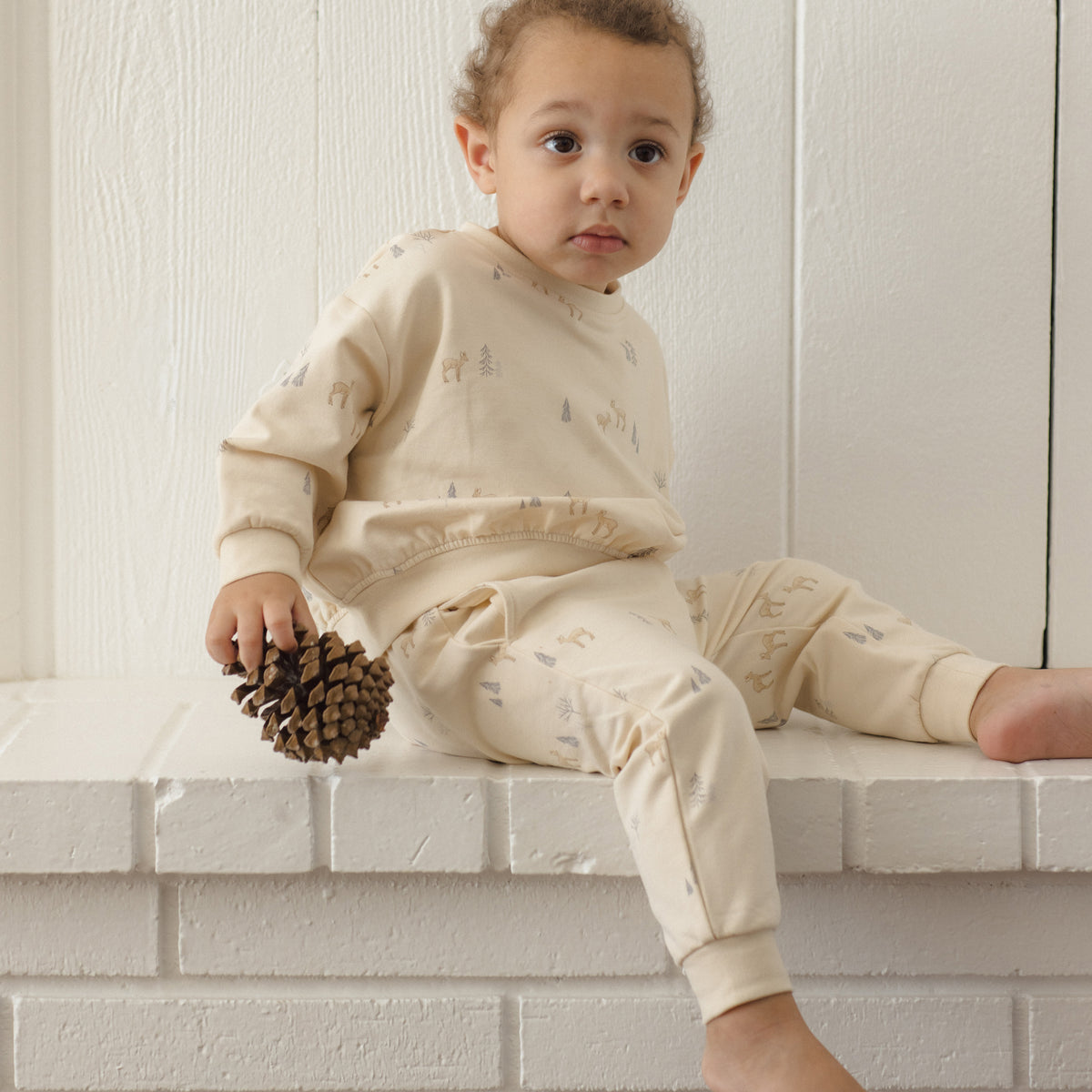 A toddler wearing a cream-colored pajama set with a subtle pine tree pattern sits on a white brick ledge holding a pinecone against a white paneled wall.