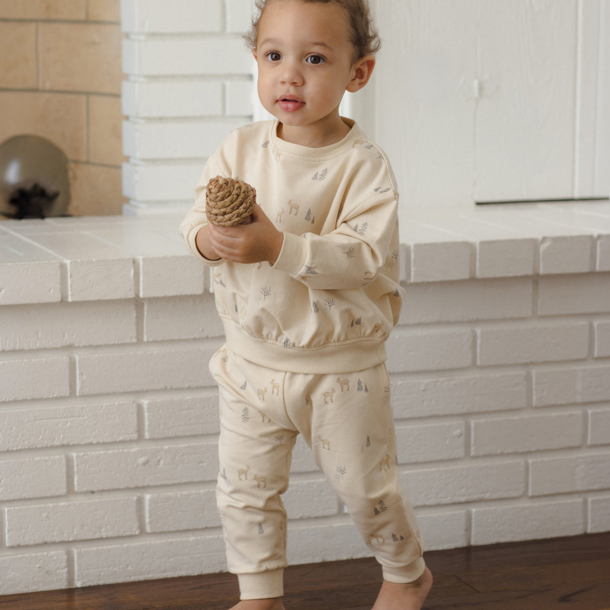 A toddler stands barefoot indoors in front of a white brick fireplace, wearing a cream-colored long-sleeve sweatshirt and matching sweatpants with a subtle allover animal print. The child is holding a small pinecone.
