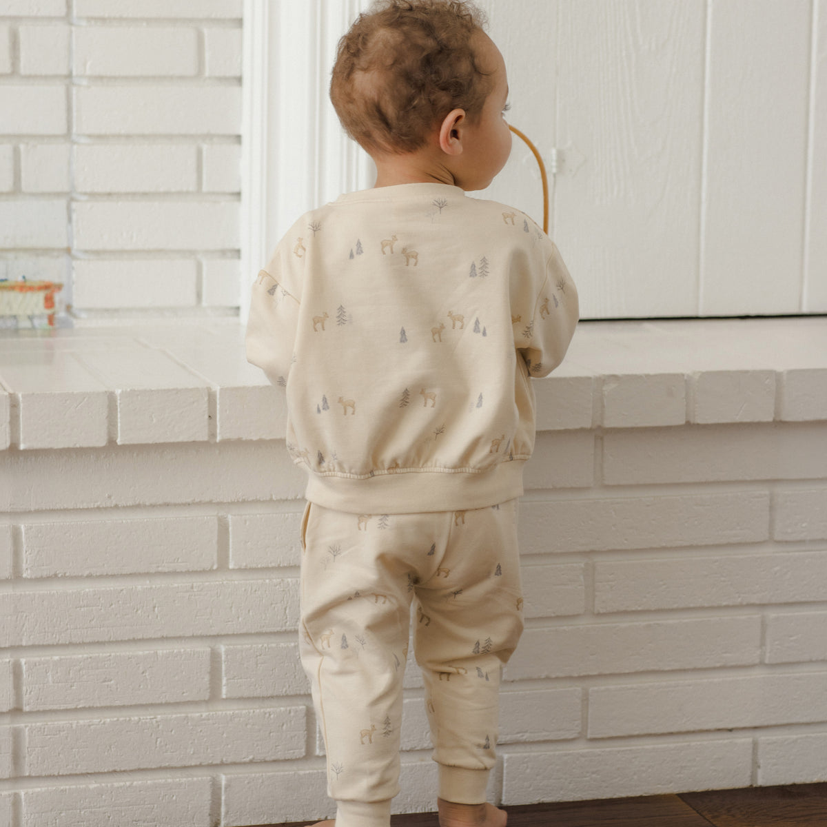 A toddler standing barefoot in front of a white brick wall, wearing a light beige long-sleeve sweatshirt and matching pants with a small repeating animal print.