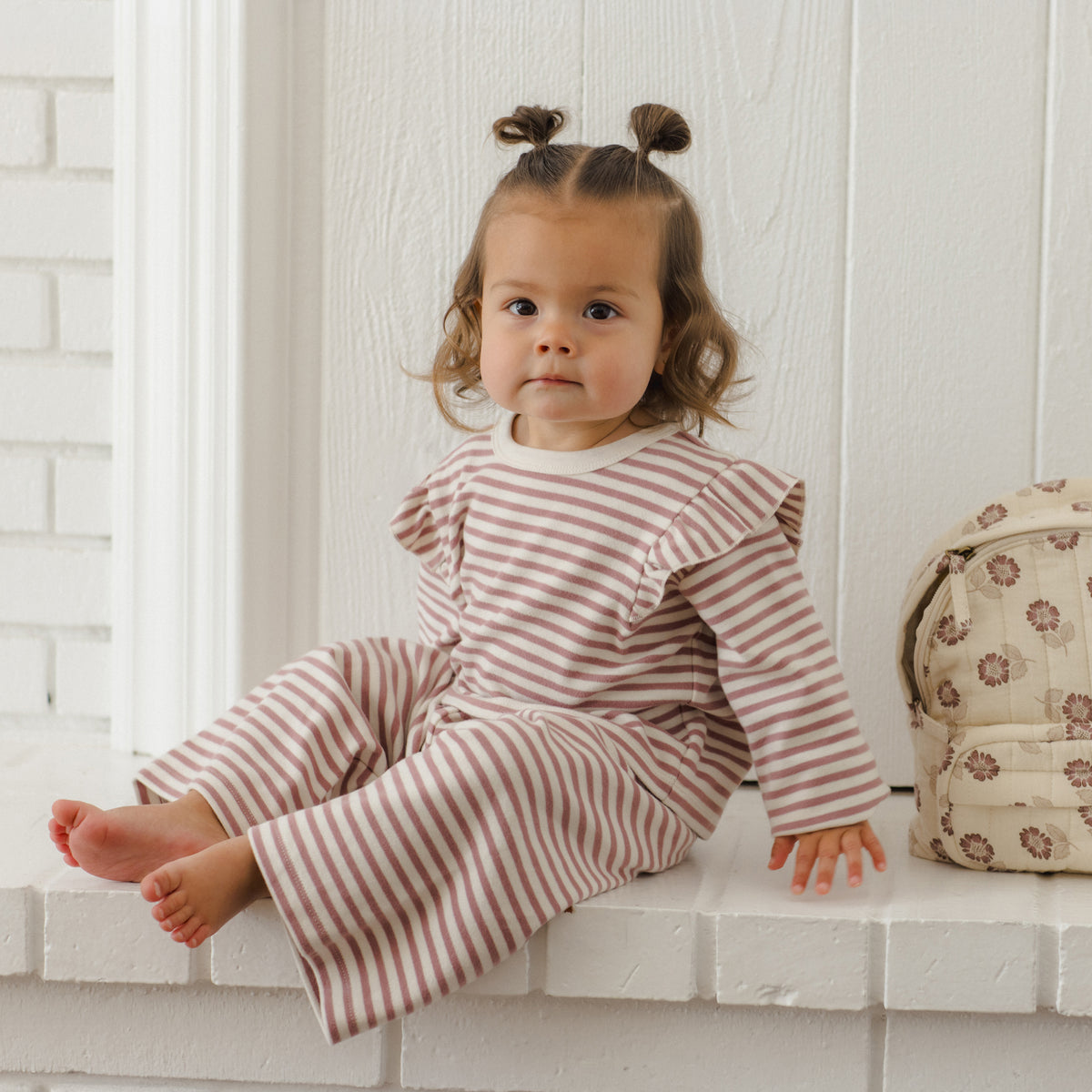 A toddler girl with two buns sits on a white brick ledge wearing a matching long-sleeve striped romper. The romper has thin reddish-brown and cream horizontal stripes.