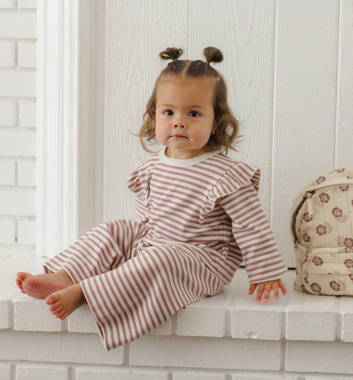 A toddler girl with two buns sits on a white brick ledge wearing a matching long-sleeve striped romper. The romper has thin reddish-brown and cream horizontal stripes.