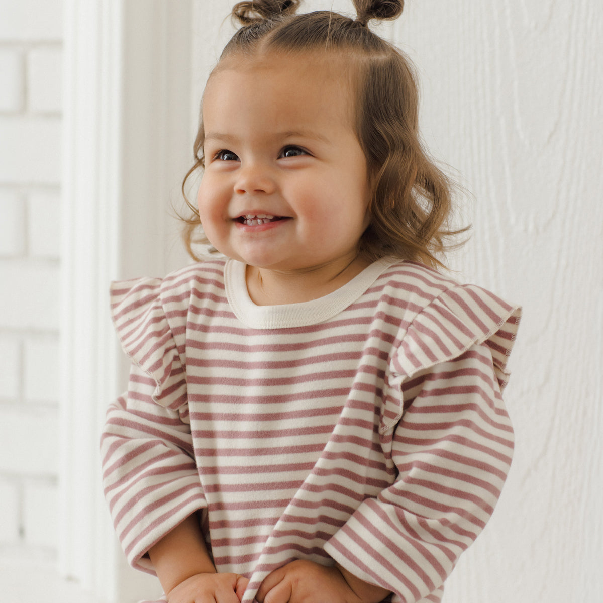 A smiling toddler with two buns sits wearing a long-sleeve ruffled shirt and matching striped pants in a soft pink and white pattern, set against a bright white interior background.