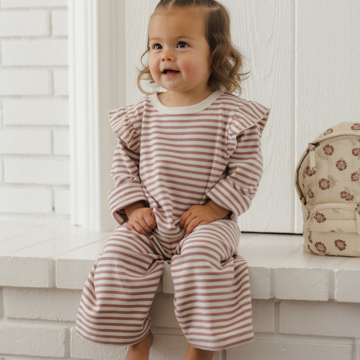 A toddler girl with two small buns sits barefoot on a white brick ledge, wearing a long-sleeve ruffled top and matching wide-leg pants in a dusty pink and white stripe pattern. A small patterned cream backpack rests on the ledge to her right.