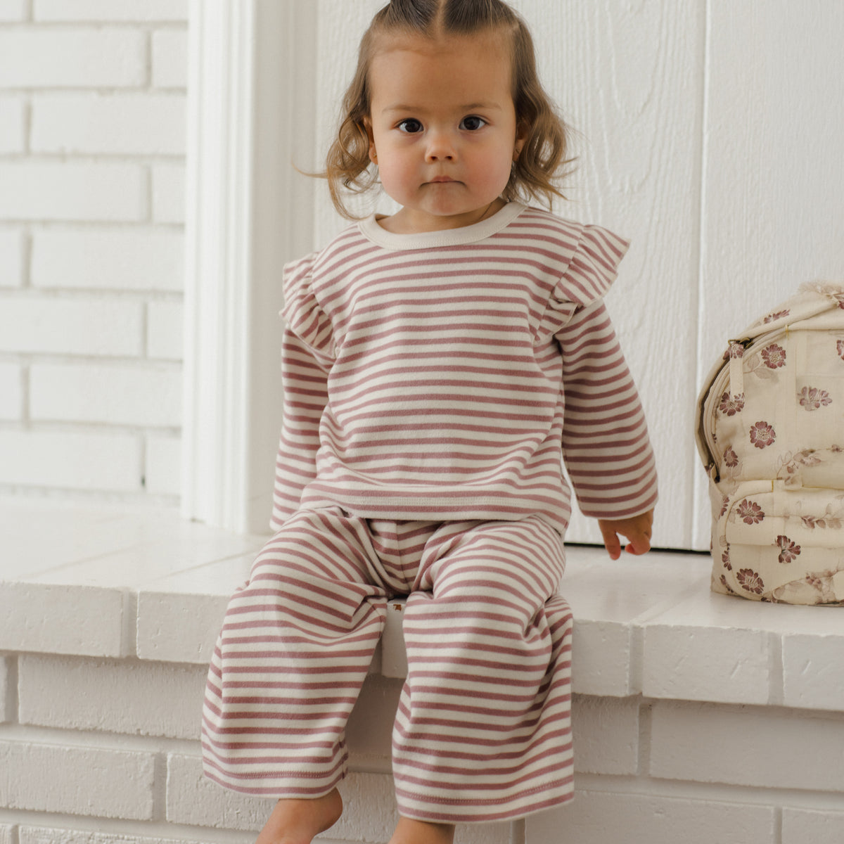 A toddler girl with two small buns sits barefoot on a white brick ledge, wearing a matching long-sleeve top and wide-leg pants set in a dusty pink and cream stripe pattern. A floral-patterned bag rests on the right side of the ledge.