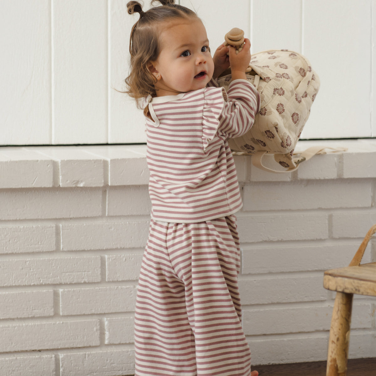 A toddler stands barefoot indoors against a white paneled wall, wearing a matching two-piece pajama set with thin brown stripes on a warm off-white background. The set includes a long-sleeve top and wide-leg pants.