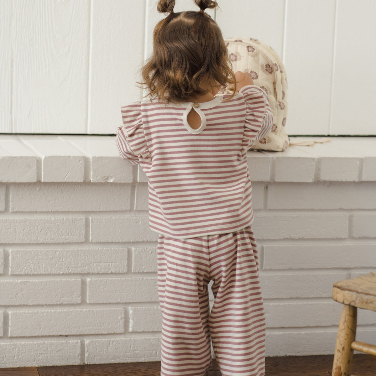 A toddler viewed from behind wearing a matching two-piece pajama set with thin pink and white horizontal stripes, featuring a long-sleeve top with a keyhole cutout and wide-leg pants. The child is standing in front of a white brick wall and white paneled wall.
