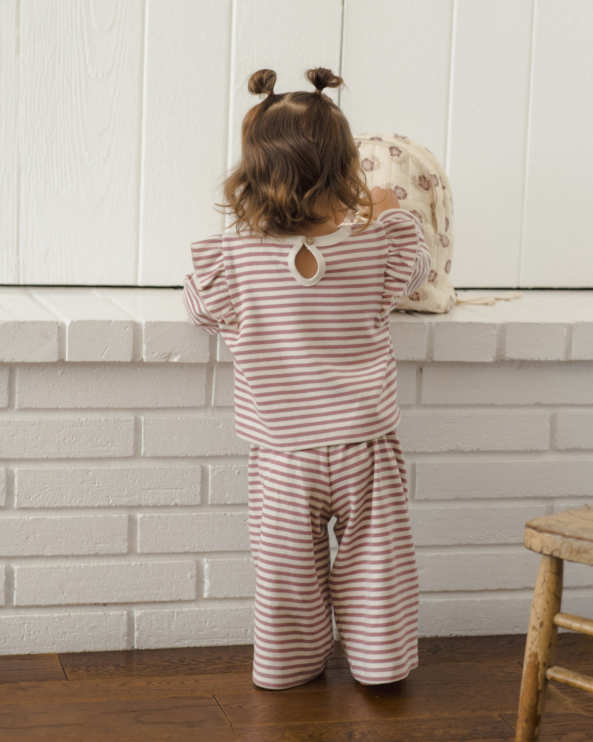A toddler viewed from behind wearing a matching two-piece pajama set with thin pink and white horizontal stripes, featuring a long-sleeve top with a keyhole cutout and wide-leg pants. The child is standing in front of a white brick wall and white paneled wall.