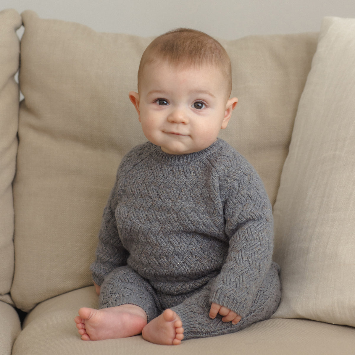 A baby sitting on a beige sofa wearing a gray cable-knit long-sleeve romper.