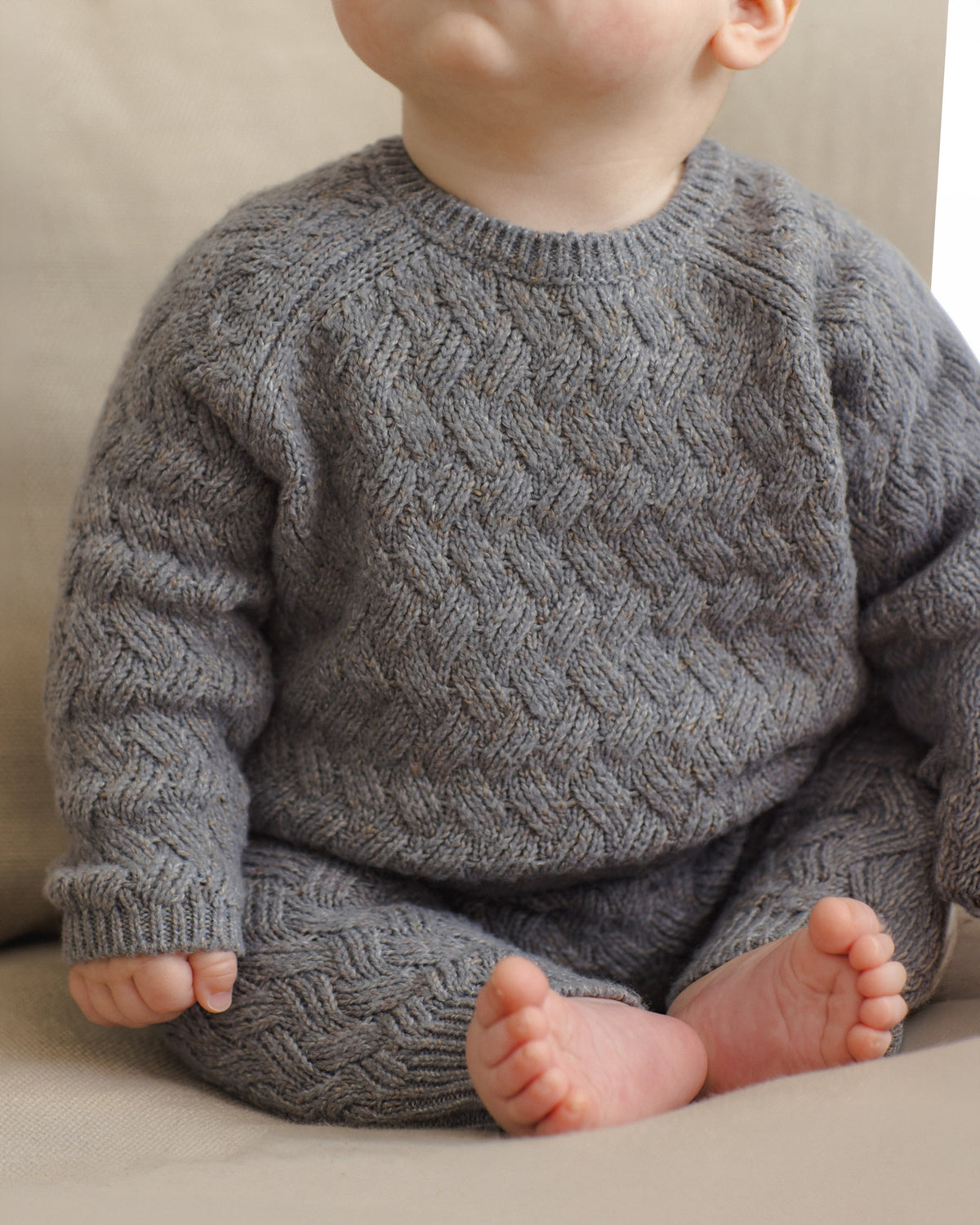 A baby wearing a gray cable-knit long-sleeve romper or sweater, sitting against a neutral beige background.