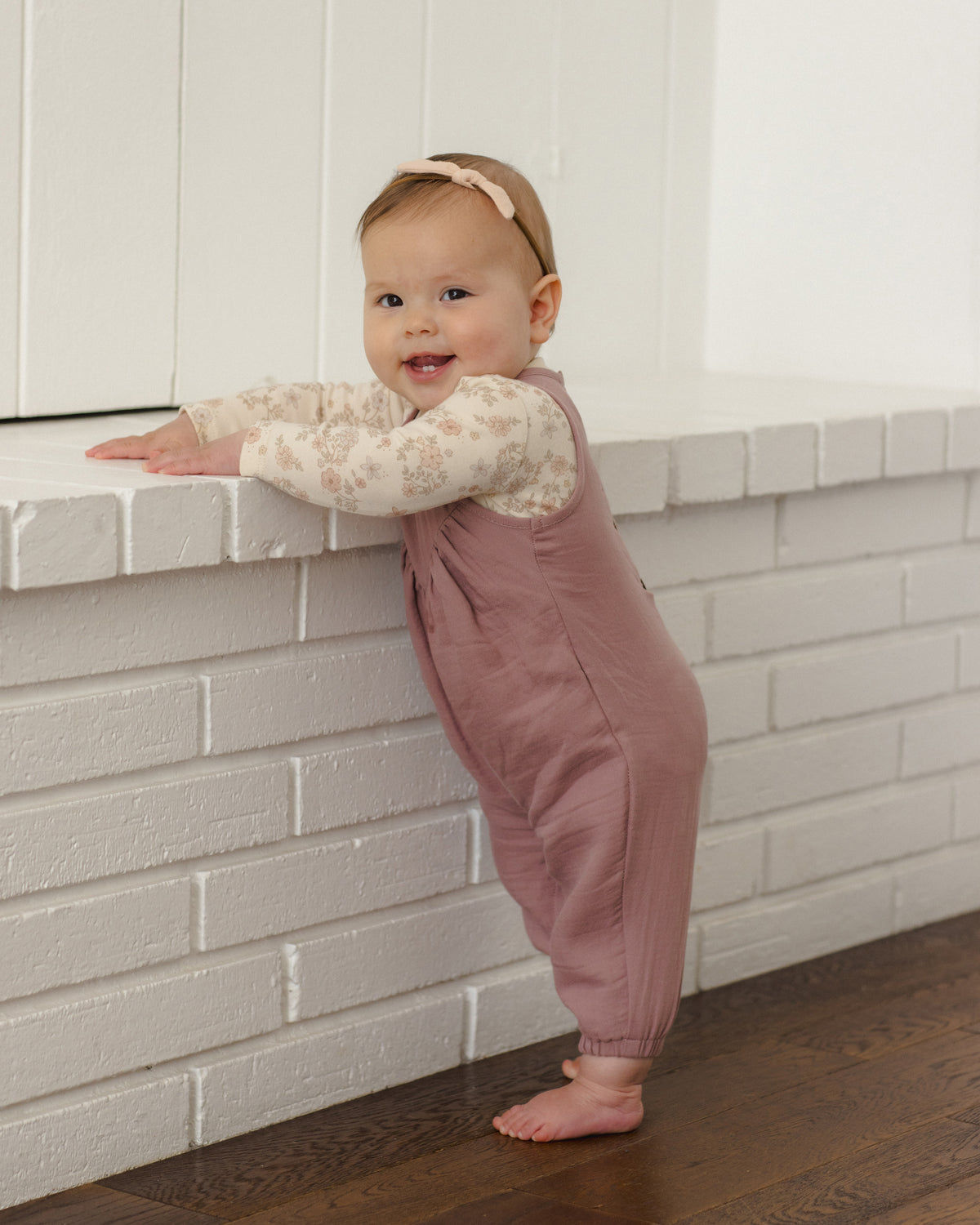 A baby standing on a dark wood floor next to a white brick fireplace, wearing a dusty rose overall jumpsuit over a cream floral long-sleeve shirt and a matching beige headband.