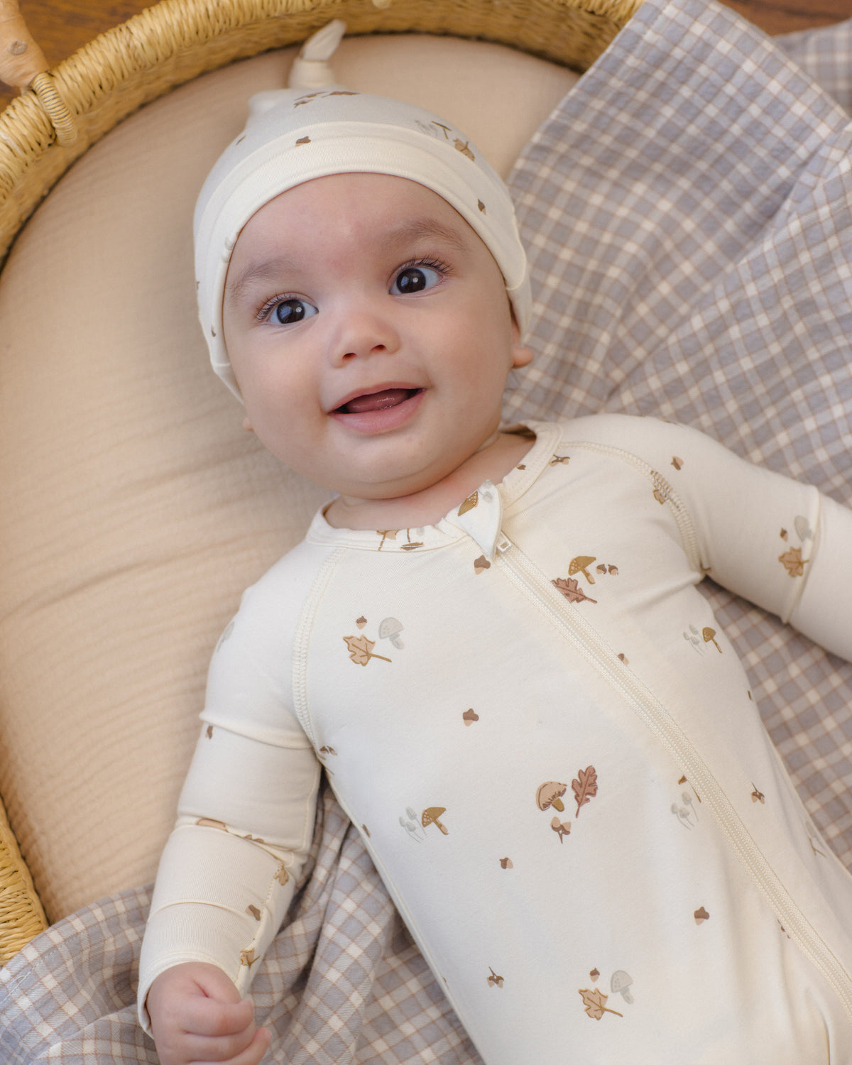 A baby lying in a woven bassinet wearing a cream-colored zip-up sleeper and matching bonnet with small brown woodland animal prints, set against a beige and gray check blanket.