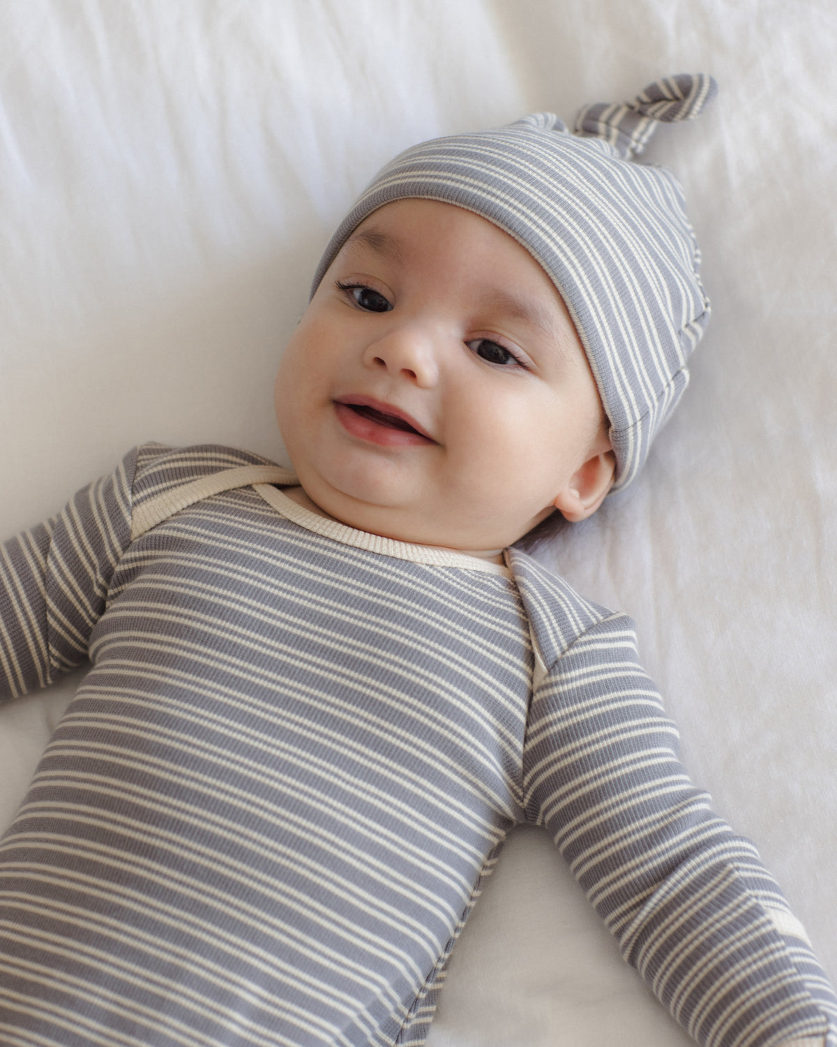 A baby lying on a white blanket, wearing a gray and white striped long-sleeve bodysuit and a matching striped beanie with a small bow on the tip.