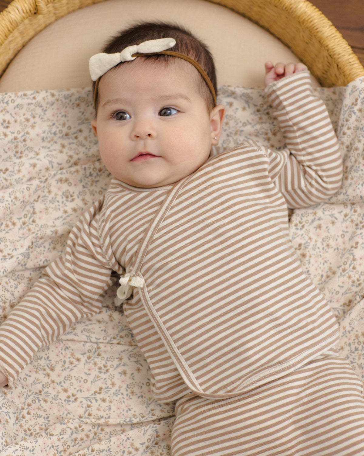A baby lying on a floral mattress inside a wicker bassinet, wearing a beige and white striped long-sleeve sleep sack and a matching white bow headband.