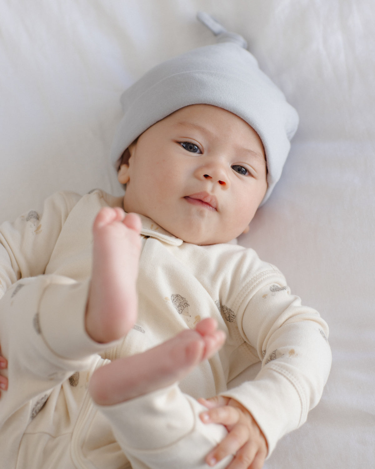 A baby lying on a white blanket, wearing a light blue knit beanie and a cream-colored long-sleeve footed onesie with small gray animal prints.