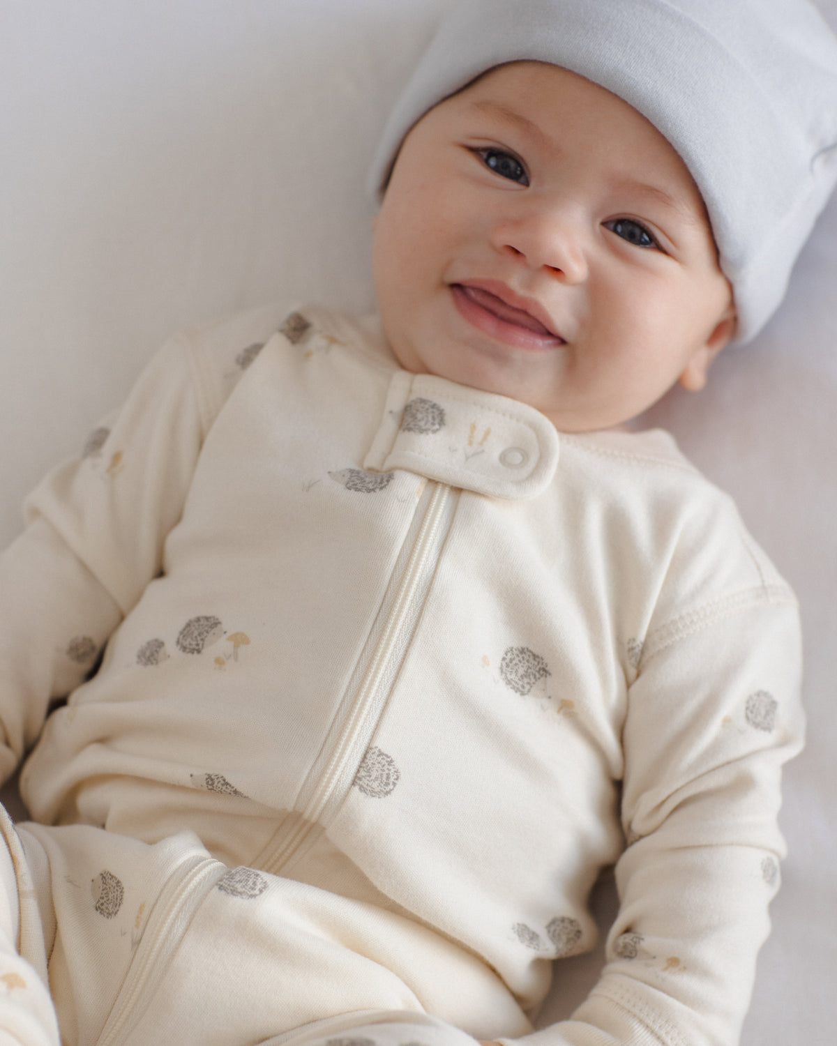A smiling baby lying down wearing a cream-colored long-sleeve zip-up sleeper with small gray and beige mushroom prints, along with a light blue beanie.