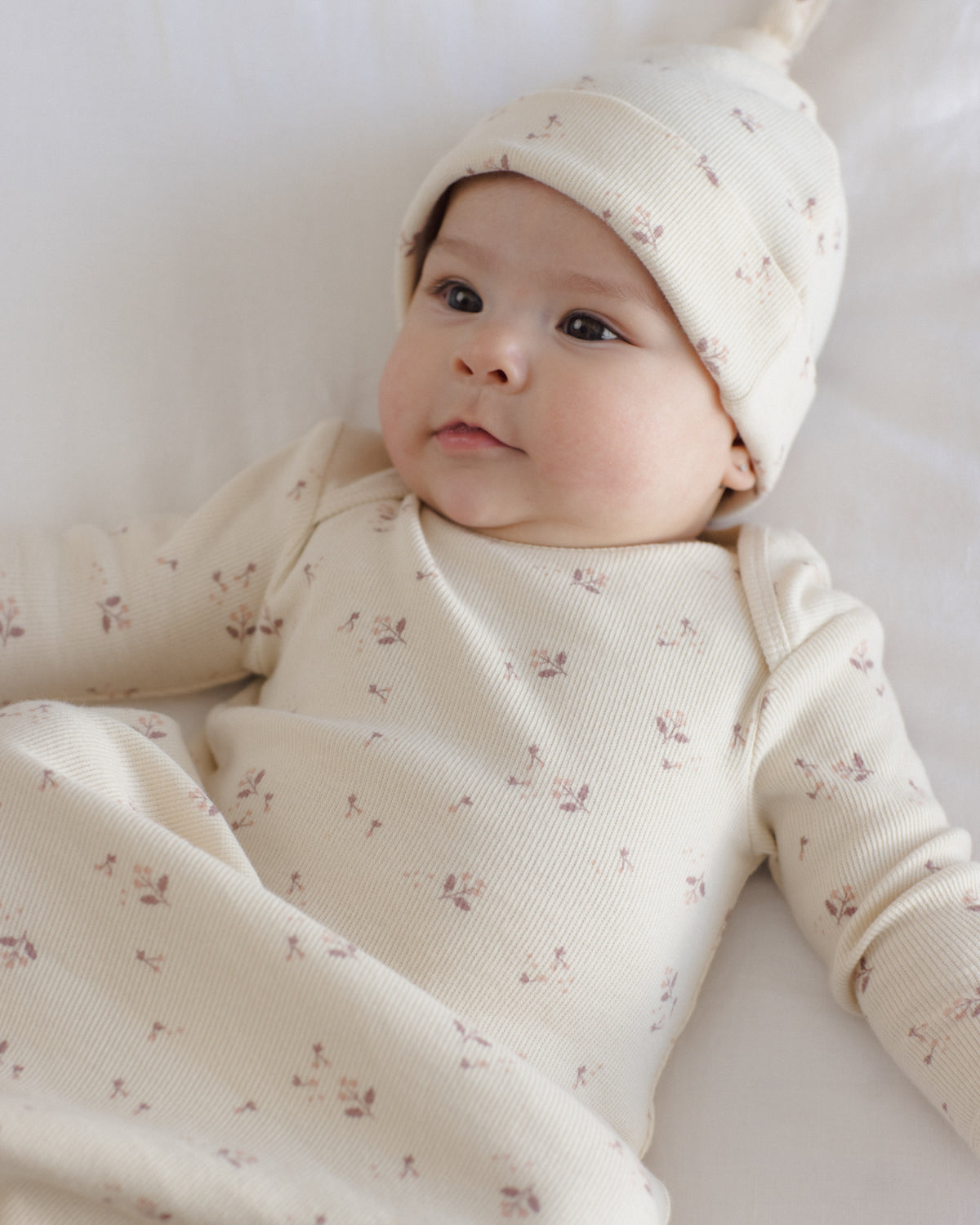 A baby lying on a white blanket, wearing a cream-colored ribbed knit one-piece outfit with a small pink floral print and a matching pointed hat.