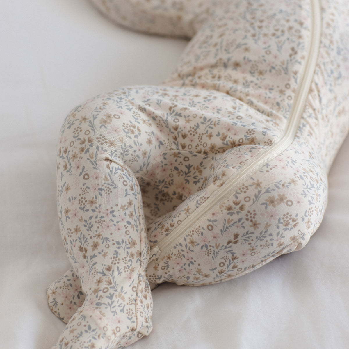 A baby lying on a white bed wearing a footed one-piece sleeper with a delicate beige, blue, and brown floral pattern and a front zipper.