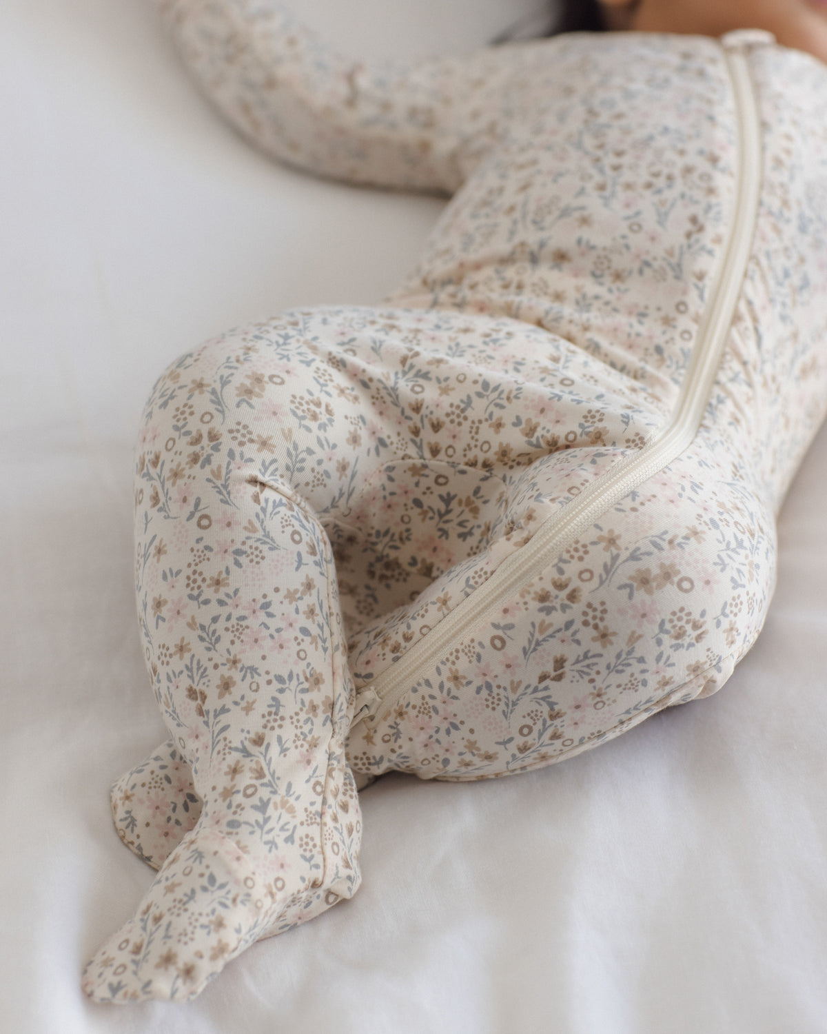 A baby lying on a white bed wearing a footed one-piece sleeper with a delicate beige, blue, and brown floral pattern and a front zipper.