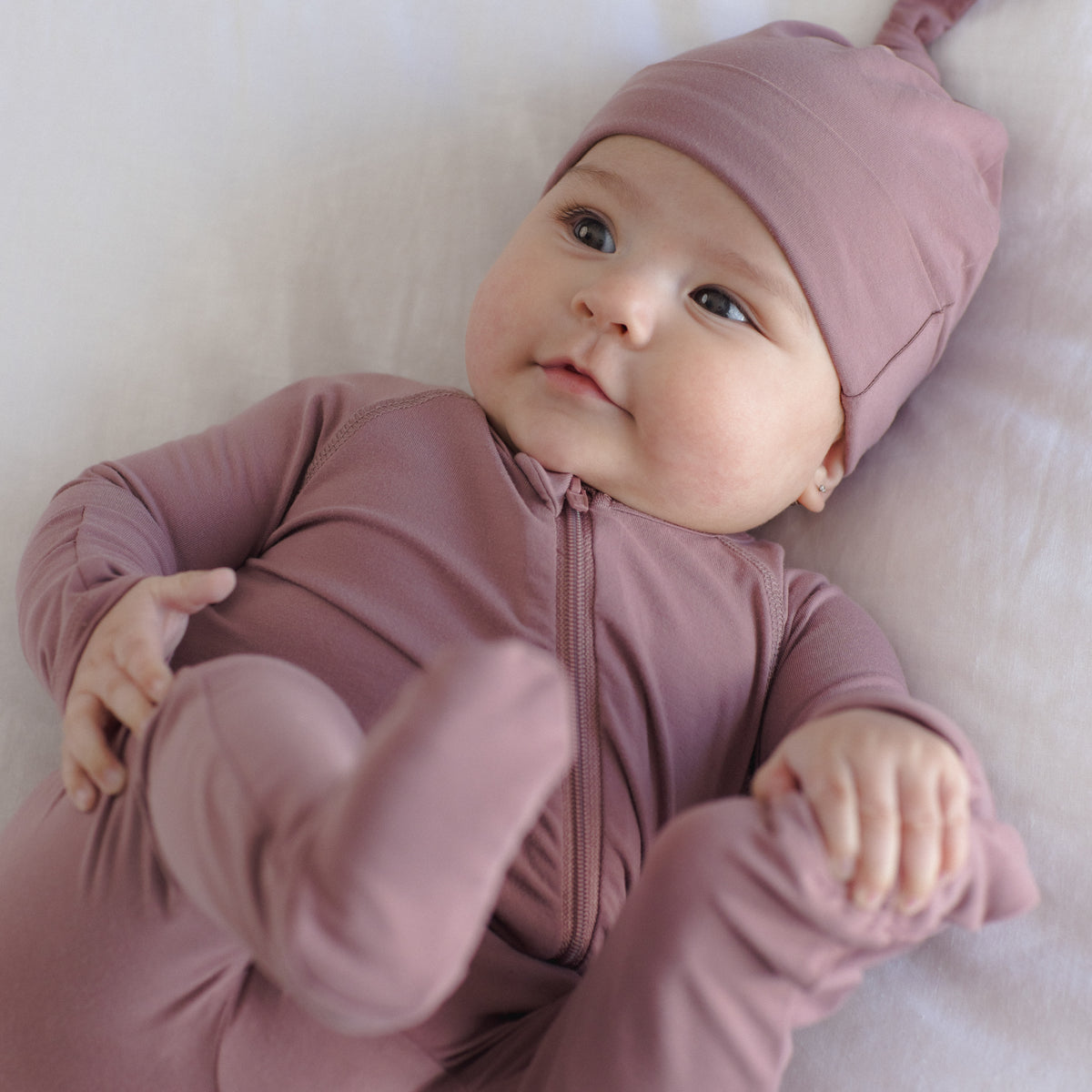 A baby lying on a white blanket, wearing a dusty mauve zip-up one-piece sleeper with a matching pointed knit hat.