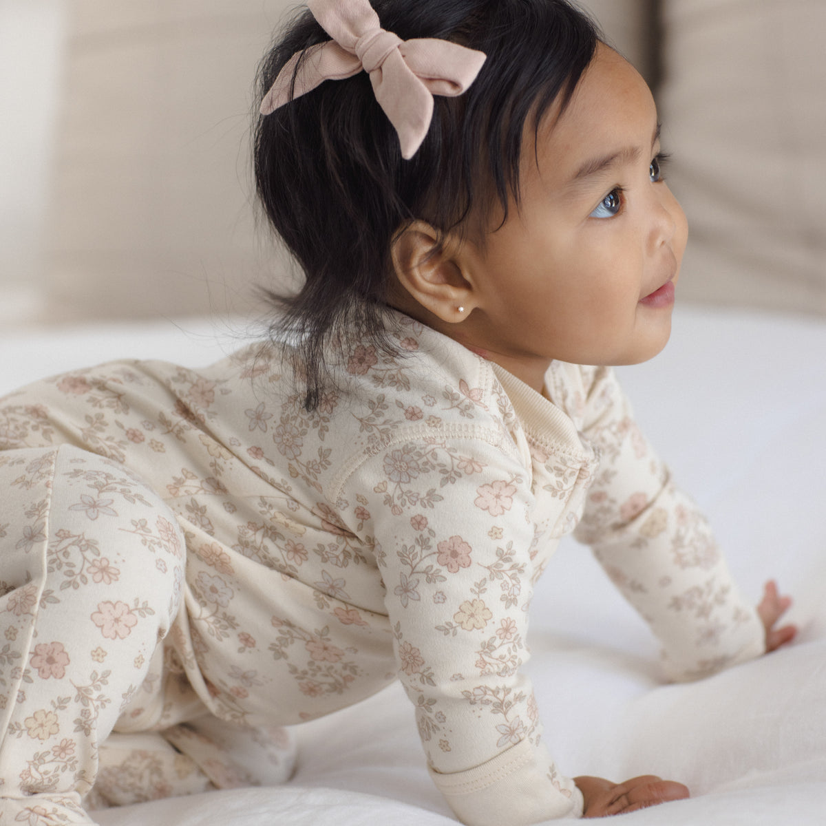 A baby crawling on a white bed wearing a cream-colored long-sleeve footed sleeper with a delicate pink floral pattern and a matching pink bow headband.