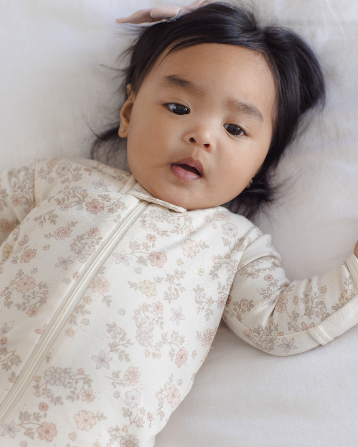 A baby lying on a white blanket, wearing a cream-colored long-sleeve sleep sack with a delicate pink and beige floral pattern.