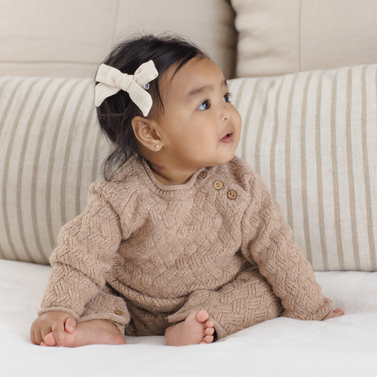 A baby wearing a beige cable-knit romper and matching hat, sitting on a white blanket in front of striped pillows.